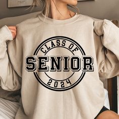 a woman sitting on a bed wearing a sweatshirt that says class of senior in black