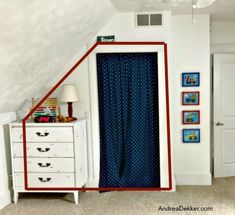 an attic bedroom with blue curtains and white dresser in the foreground, and pictures on the wall