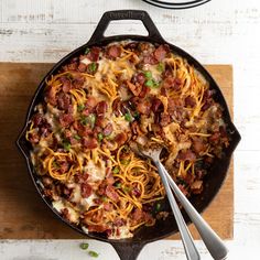 a skillet filled with spaghetti and bacon on top of a wooden cutting board next to a pot