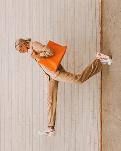 a woman standing on one leg and holding an orange bag in her other hand while leaning against a wall