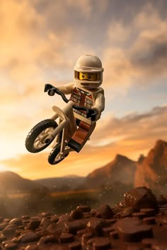 a lego man riding a dirt bike in the air over rocks and grass at sunset