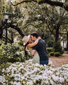 a man and woman kissing in front of white flowers with trees behind them at the park