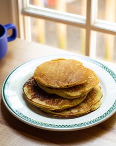 three pancakes on a white plate next to a blue coffee mug and window sill