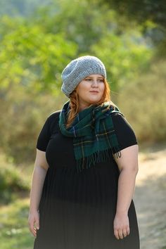 a woman wearing a black dress and a green plaid scarf is standing on a dirt road