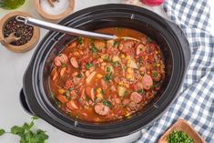 a crock pot filled with sausage and beans on top of a table next to vegetables