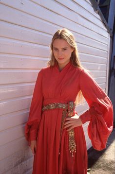 a woman in a red dress leaning against a white wall with her hands on her hips