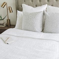 a bed with white sheets and pillows on top of it next to a lamp, books and a potted plant