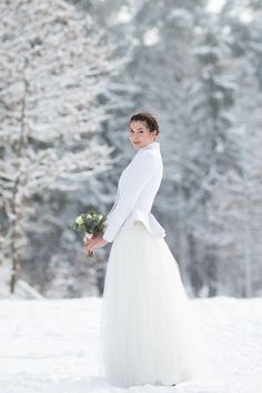 a woman standing in the snow wearing a white wedding dress and holding a flower bouquet