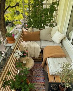 an outdoor balcony with lots of plants and pillows on the couches, potted trees in the background