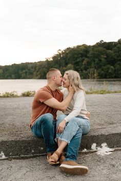 a man and woman sitting next to each other on concrete near water with trees in the background