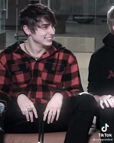 two young men sitting next to each other in front of a glass wall, one wearing a red and black checkered shirt