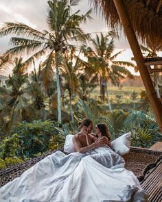 two people sitting on a hammock in the middle of palm trees and grass