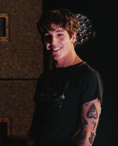 a smiling young man with tattoos on his arm and chest, standing in front of a wall