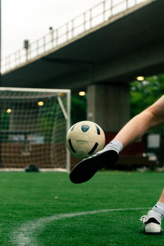 a man kicking a soccer ball on top of a green field with a bridge in the background