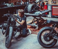 a man sitting on the ground next to two motorcycles in a garage with other men working on them