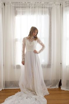 a woman is standing in front of a window wearing a wedding dress with long sleeves