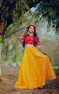 a woman in a yellow skirt and red top standing under a tree with her hands on her head