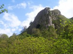 a mountain with a cave in the middle surrounded by green trees and blue cloudy sky