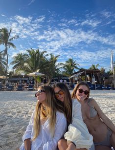 three women sitting on the beach with palm trees in the background