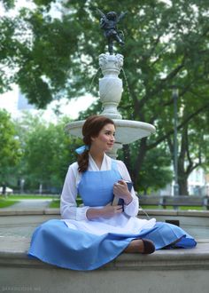 a woman sitting on the ground in front of a fountain wearing an apron and dress