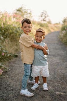 two young boys are hugging each other on a dirt road in front of some flowers