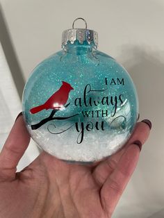 a hand holding a glass ornament with a red bird on it that says i am always with you