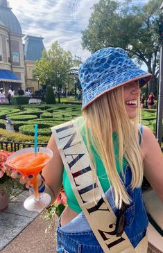 a woman in overalls and a blue hat is holding a drink at an amusement park