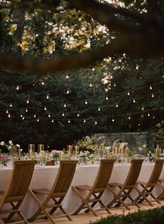 a long table with chairs and lights hanging from the trees in front of it is surrounded by greenery