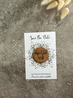 a wooden save the date magnet sitting on top of a stone floor next to some dried flowers