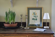 a wooden table topped with a potted plant next to a lamp and framed painting