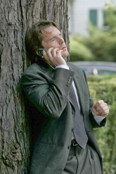 a man in a suit leaning against a tree talking on a cell phone while holding his hand up to his ear