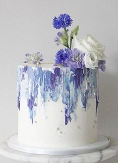 a white cake with blue and purple flowers on top is sitting on a marble stand