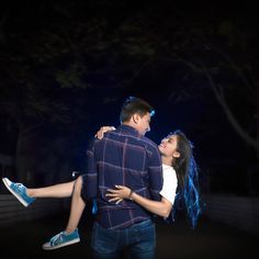 a man holding a woman in his arms at night with the lights on behind him