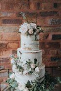 a three tiered cake with flowers and greenery on the top is sitting in front of a brick wall