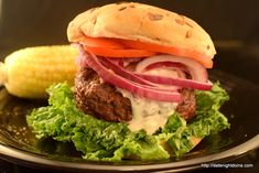 a hamburger with lettuce, tomato and onion on a black plate next to corn