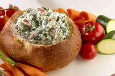 a bowl filled with spinach dip surrounded by carrots, cucumbers and tomatoes