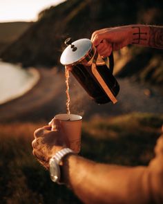 a person pouring coffee into a cup on the side of a hill next to a body of water