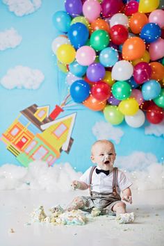 a baby sitting on the ground with balloons in the air behind him and his mouth wide open