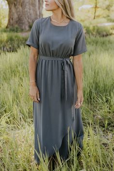 a woman standing in tall grass wearing a gray dress with short sleeves and a belt