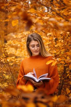 a woman in an orange sweater is reading a book while surrounded by trees with yellow leaves