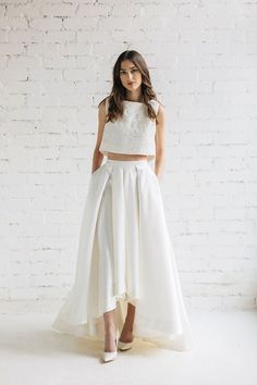 a woman standing in front of a brick wall wearing a white skirt and crop top