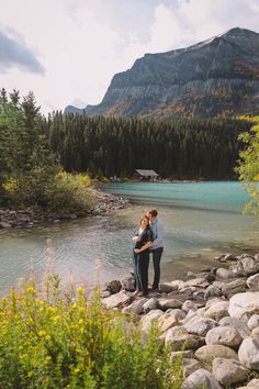 a man and woman standing next to each other on rocks near water with mountains in the background