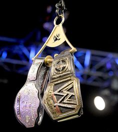 a pair of wrestling belts hanging from a metal hook on a black background with lights in the background