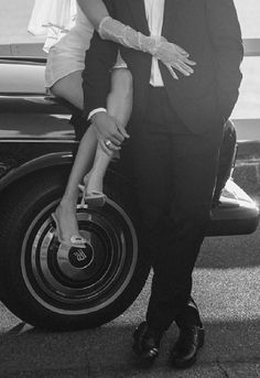 a man and woman standing next to each other in front of a car with their hands on the wheel