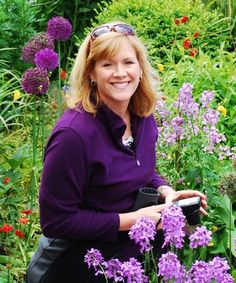 a woman sitting on a bench in front of purple flowers and green grass, smiling at the camera