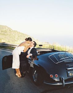 a bride and groom kissing in front of a black sports car on the side of the road