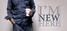 a man holding a coffee cup standing in front of a wall that says i'm new here