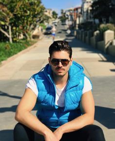 a man sitting on the sidewalk with his feet crossed wearing sunglasses and a blue vest