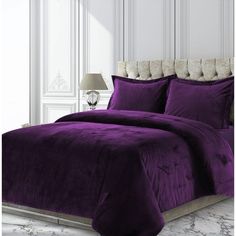 a bed with purple comforter and pillows in a white room next to a lamp