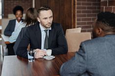 a man sitting at a table with a cup of coffee in front of him and two other people behind him